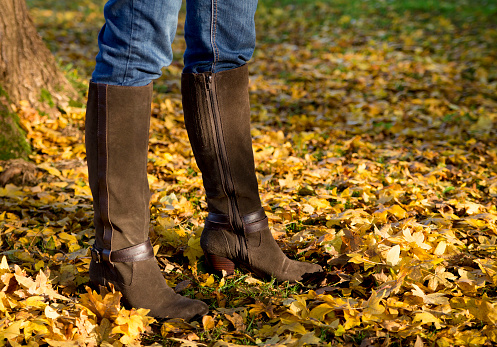 A woman's legs wearing brown leather knee high boots standing in an outdoor scene among golden autumn leaves.