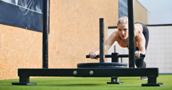 Muscular and strong young female pushing the prowler exercise equipment on artificial grass turf. Fit woman exercising at gym gym.