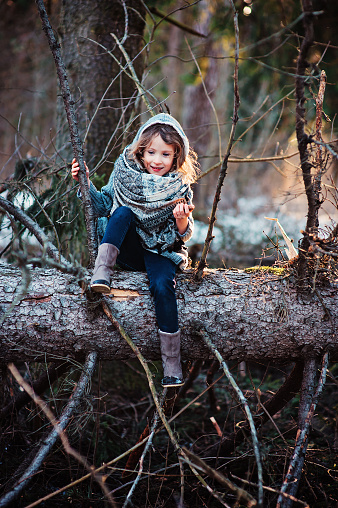 child girl in headscarf sitting and playing on huge old pine tree on winter walk