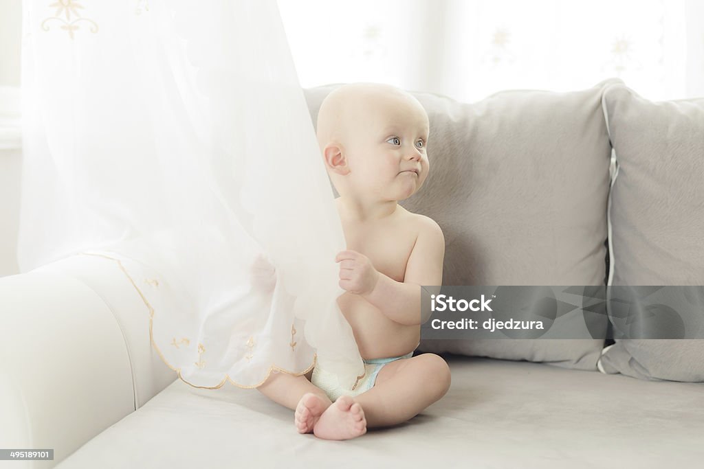 aby sitting on the sofa in  living room baby sitting on the sofa in the living room and hides behind a curtain. dressed only in a diaper 6-11 Months Stock Photo