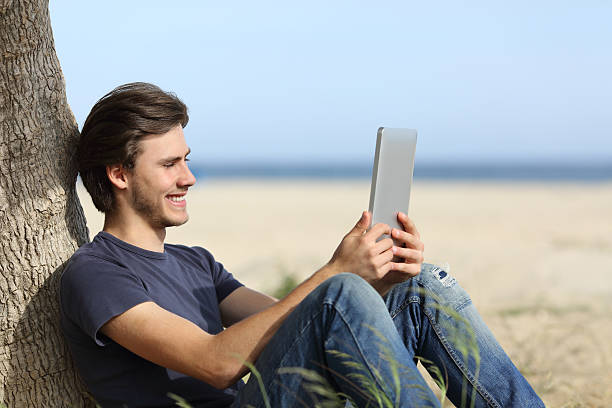 feliz homem lendo um tablet reader sentado na praia - reading beach e reader men - fotografias e filmes do acervo