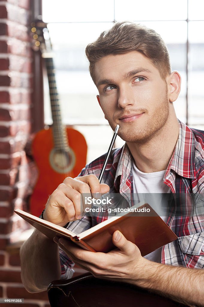 Creative inside his soul. Handsome young man writing something in note pad and touching his chin with pen while acoustic guitar laying in the background Acoustic Guitar Stock Photo