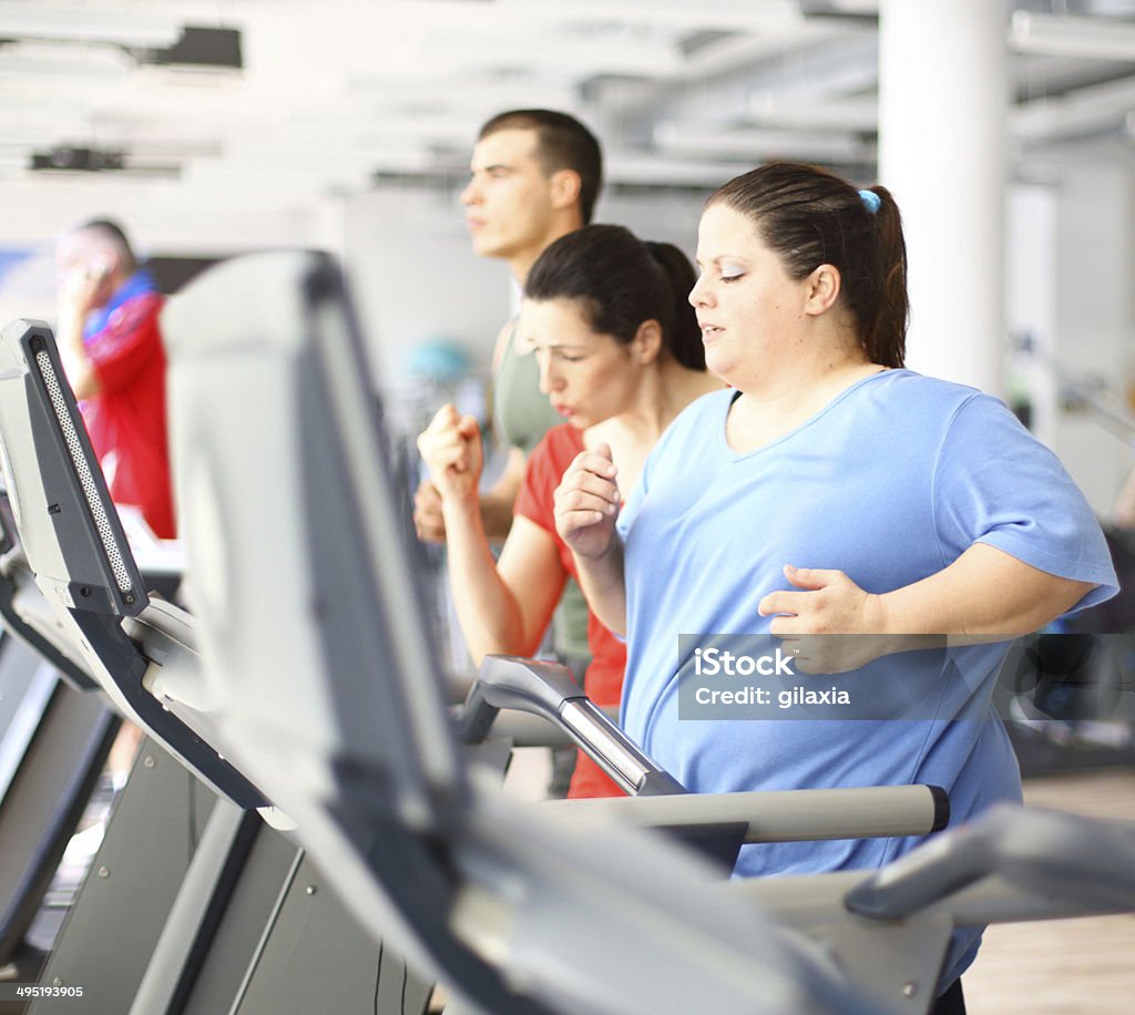 Overweight woman exercising on a treadmill. Overweight woman doing cardio workout on a treadmill with her coach assisting.She's doing light jogging and wearing blue t-shirt.Her coach is strongly supporting her in red tank top.There are several people in background exercising,out of focus. Overweight Stock Photo