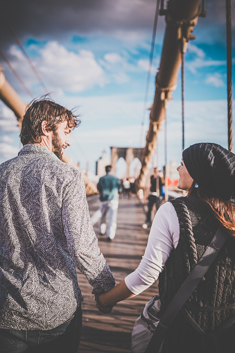 DSLR picture of a couple in love walking hand in hand on the famous Brooklyn Bridge during a beautiful sunny autumn day. They are looking at each other and are happy and smiling. 