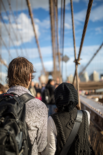 DSLR picture of a couple in love walking hand in hand on the famous Brooklyn Bridge during a beautiful sunny autumn day. The city buildings of New York are visible in the background. 