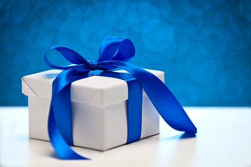 White gift box tied up with blue ribbon bow sitting on a white table against blue defocused lights. Predominant colors are blue and white. DSRL studio photo taken with Canon EOS 5D Mk II and Canon EF 70-200mm f/2.8L IS II USM Telephoto Zoom Lens