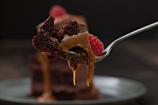 An extreme close up horizontal photograph of a piece of chocolate fudge cake on a fork dripping golden brown caramel sauce, isolated on black.