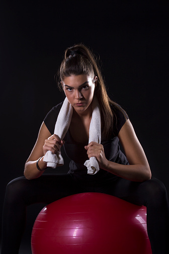 Long dark hair beautiful woman sitting on red pilates ball and holding white towel over her neck and looking at the camera