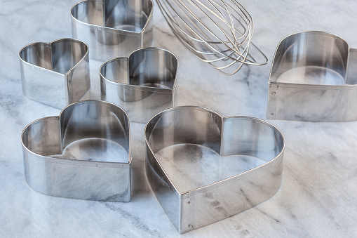 Studio photo of heart shaped stainless steel cookie cutters and whisk on light colored marble. Selective focus on tops of metal shapes.