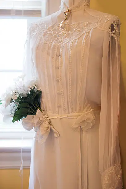 Partial view of the bodice of a white dress with lace, beading, pearls, covered buttons and a pearl drop brooch from a high neckline and a sleeve wrapped around an arrangement of artificial flowers