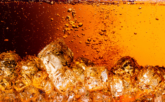 Stock photo showing close-up view of some ice cubes being dropped into an American diner style glass of fizzy cola, causing splashing and a secondary splash of water droplets.