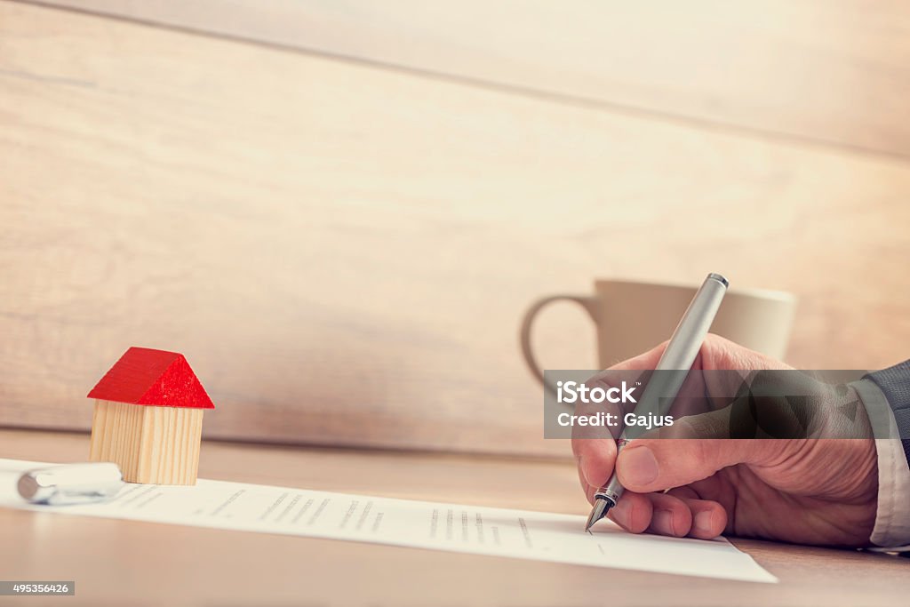 Closeup of male hand signing insurance papers, contract of house Closeup of male hand signing insurance papers, contract of house sale or mortgage documents with fountain pen, wooden toy house sitting on paperwork. 2015 Stock Photo