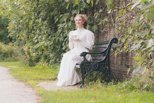 Posh victorian woman having cup of tea on garden seat.