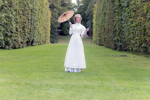 Victorian fashion woman holding parasol standing on lawn with tall hedge.