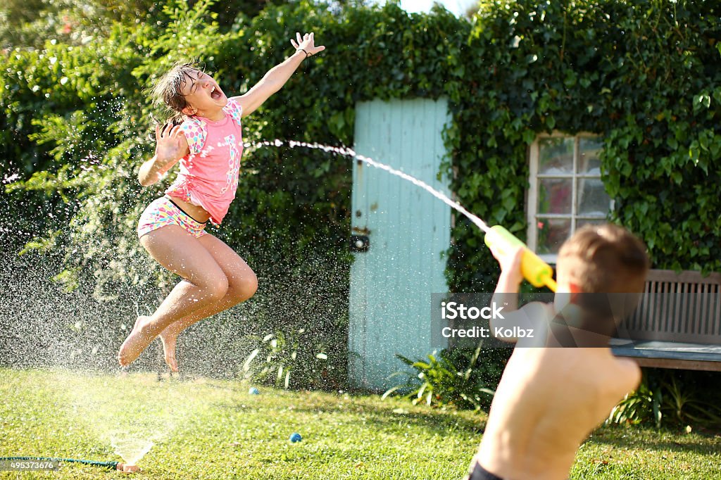 Water Fight A young boy shoots his sister with a water gun as she jumps in a garden hose sprinkler in the back yard on a hot summer afternoon Child Stock Photo