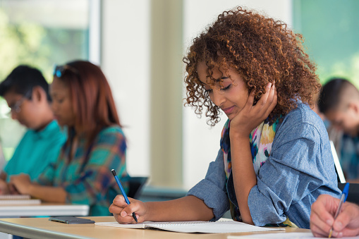 Teenage African American high school or college student is studying paper and writing while taking a test in modern classroom. Girl has curly hair and is wearing trendy casual clothing. She is sitting at a desk with diverse classmates in background.