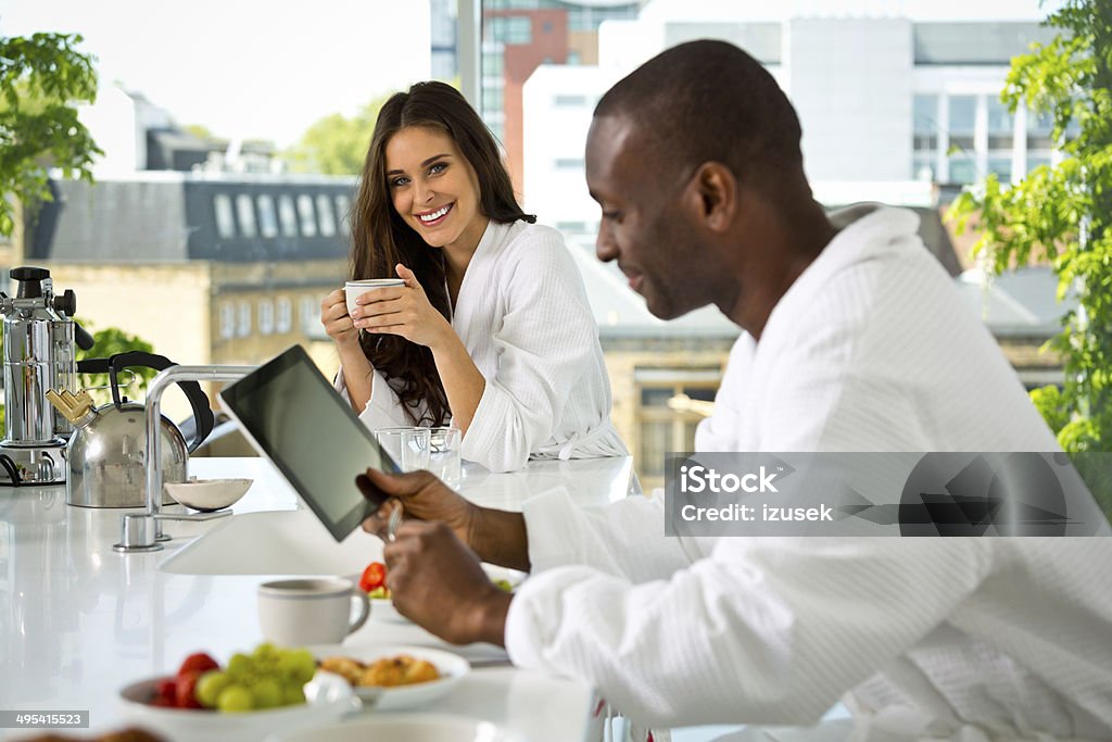 Happy morning Afro american man and beautiful brunette wearing white bathrobes having breakfast in their apartment, woman drinking coffee and man using a digital tablet. 30-39 Years Stock Photo