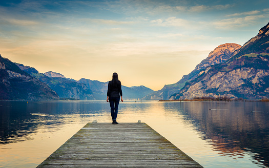 Epic mountain landscape. Female figure on a wooden pier. Lake at sunset.