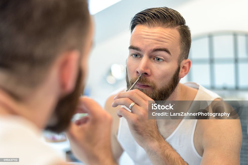 Man plucking his nasal hairs Handsome bearded young man standing plucking his nasal hairs in front of the mirror in the bathroom during his daily grooming Nose Stock Photo