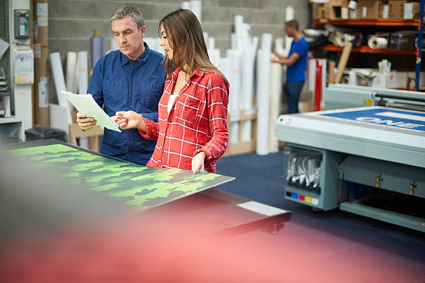 woman working at a signage company A young woman is working on a digital printing machine . her boss is explaining the machine or the job that she is doing . In the background rolls of vinyl can be seen , and a young co-worker is cutting out some vinyl lettering digital printing house stock pictures, royalty-free photos & images