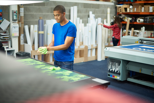 A young man is working on a digital printing machine and putting on his gloves before handling the print for inspection .  In the background rolls of vinyl can be seen , and a young co-worker is cutting out some vinyl lettering   