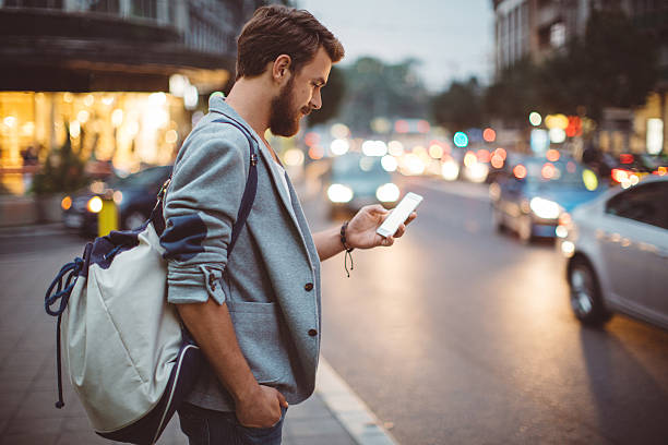 Young man on the streets of big city. Young man on the street of big city , waiting taxi, chacking his smartphone, for news or new messages.  Or looking for map instructions.  Carry backpack on one shoulder.  Dusk time.  Casual dressed. Street lights in background. taxi photos stock pictures, royalty-free photos & images