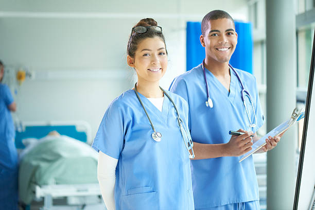 two young nurses on the ward a young male and female nurse stand proudly on the ward and look to camera . In the background a patient is being attended by a nurse and their relative . female nurse stock pictures, royalty-free photos & images