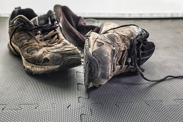 A grubby baseball cap on the health club gym flexible rubber floor mat is behind a pair of well-used athletic running sneaker shoes covered in dried mud.