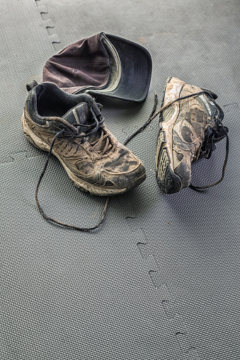 A grubby baseball cap on the health club gym flexible rubber floor mat next to a pair of well-used athletic running sneaker shoes covered in dried mud.