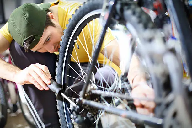 Early 30's man repairing his bike.He's tightening screws on rear derailer with a screwdriver.He's kneeling next to rear wheel,wearing yellow t-shirt and dark green baseball hat pointed backwards.