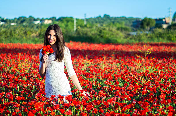 Beautiful happy smiling woman with long hair feeling freedom walking at beautiful red flowers field enjoying the stormy weather and bright sunset above it