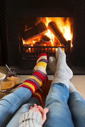 Two people are sitting with their legs infront of a fire holding hands. They are wearing wooly socks and jeans, and have one foot crossed over the other. They are sitting with their legs stretched out in a lounge. There is toast and jam on the table.