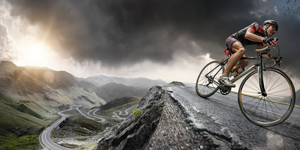 Wide angle close up image of a cyclist riding on a the peak of a road winding through mountains in a generic location, under a dramatic stormy evening sky at sunset. The bike is generic and cyclist is wearing an unbranded cycling suit, helmet and sunglasses. With intentional lighting effects. 