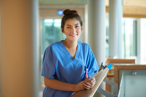 a young  female nurse stands proudly on the hospital corridor . She is wearing scrubs and holding a medical records clipboard .
