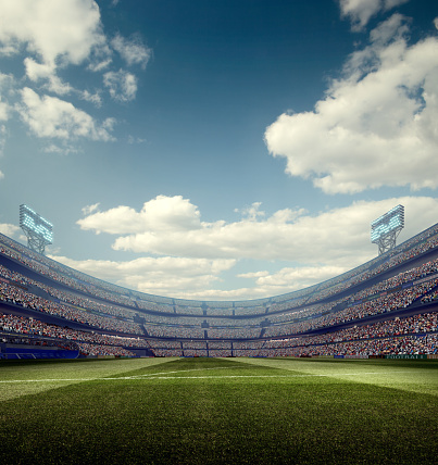 A wide angle panoramic image of a outdoor soccer stadium outdoor stadium or arena full of spectators under sunny sky. The image has depth of field with the focus on the foreground part of the pitch. 