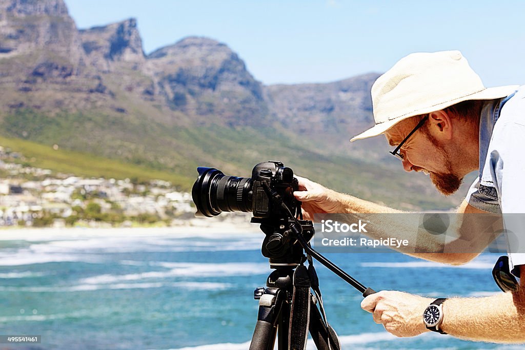 Photographer setting up shot of Camps Bay beach, Cape Town A sunhatted male photographer leans over his camera on a tripod, setting up a shot of Camps Bay beach with the Twelve Apostles mountains in the background. Another super sunny day in Cape Town! Photographer Stock Photo