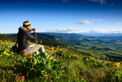 Traveler sitting in a meadow full of flowers photographing view of mountain range with Mt Hood in Cascade Range, Oregon, United States