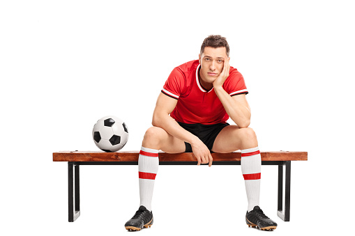 Sad young football player sitting on a wooden bench and looking at the camera isolated on white background