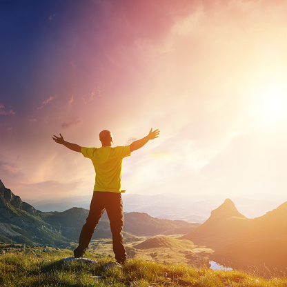 Freedom. Spirituality concept. A man standing on top of a mountain against bright sun, arms outstretched, a mountain valley on the background.