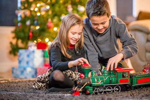 A brother and sister are sitting by the Christmas tree playing with a toy train together.