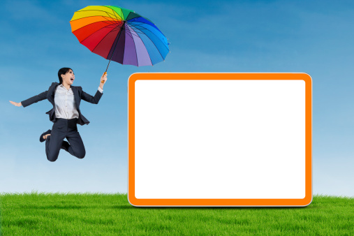 Excited businesswoman holding umbrella and jumping with blank board on the meadow