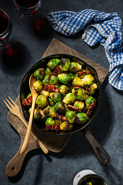 High resolution, digital capture of a cast iron skillet full of pan-seared Brussels sprouts with thick-cut, Char Siu glazed bacon . Skillet sits on a wooden cutting board, with wooden serving utensils, and a kitchen towel, plate and glass of red wine are inset. Lighting is dramatic and restrained.