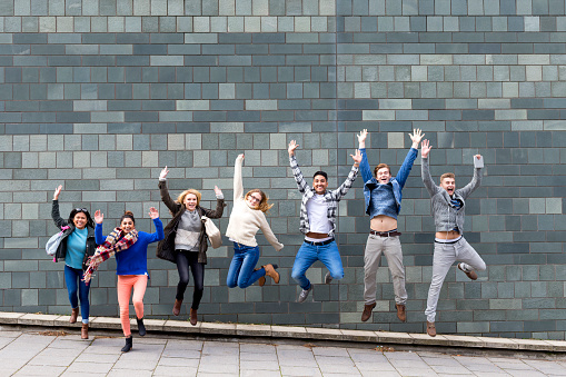 Group of young students jumping in the air in celebration. A large grey wall of their college building is behind them.