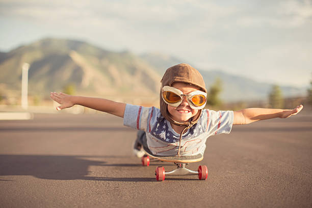 Young Boy Imagines Flying On Skateboard A young boy wearing flying goggles and flight cap outstretches his arms to attempt flying while he rides on his skateboard. He has a large smile across his face as he is imagining taking off. piloting photos stock pictures, royalty-free photos & images