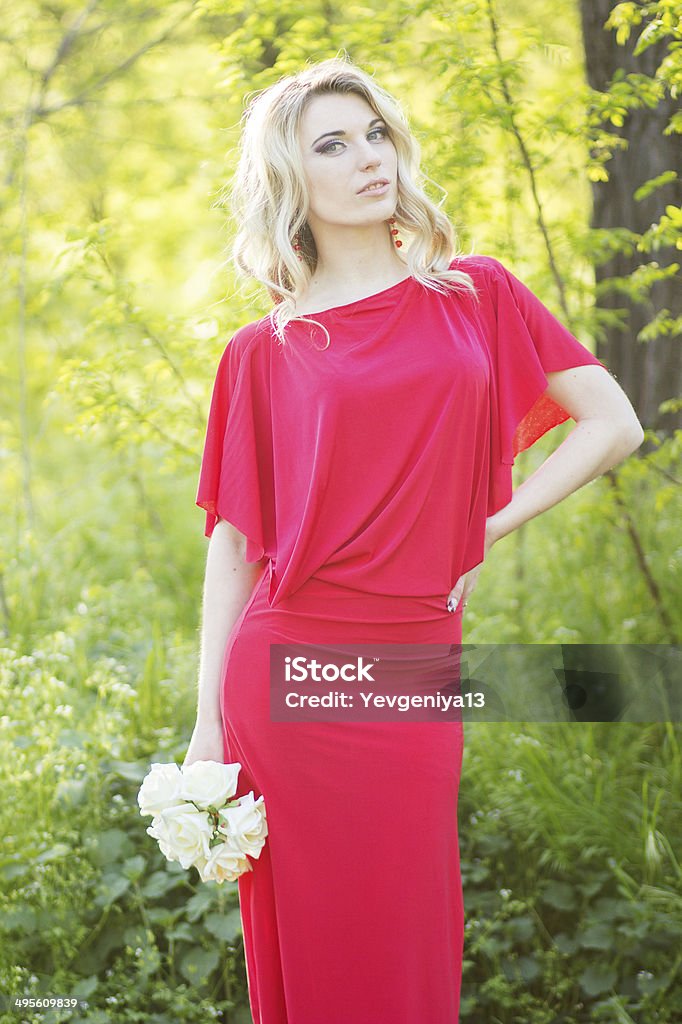 Portrait of a beautiful young blonde woman Portrait of a beautiful young blonde woman in the image of the bride. A girl wearing a red evening dress and in her hands she holds a bouquet of roses Adult Stock Photo