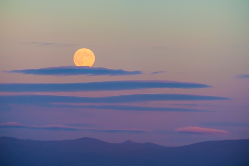 Harvest moon above the Cascade Mountain range.  The setting sun adds pastel colors to the sky while whispy clouds drift by.