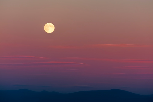 Harvest moon above the Cascade Mountain range.  The setting sun adds pastel colors to the sky while whispy clouds drift by.