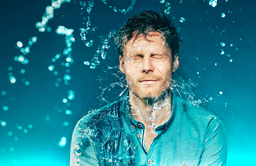Man closes his eyes, as he is hit with a bucket of water. The water splashes from his face. The photo is shot at high speed sync.