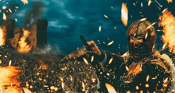 A warrior in old historic clothes - could be a knight or a viking - wears a helment and attacks with a sword. Fire and sparkles fly through the air as the man screams and gets ready to defeat an opponent. A castle is going up in flames in the background.