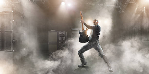 Rock Star Male rock star musician in dramatic pose wearing black clothes and leather trousers, holding up generic guitar and singing with mouth open and head back.  The guitarist is performing on a generic small indoor floodlit stage in music venue, full of sound equipment and speaker stack with haze and dry ice smoke effects.  guitarist stock pictures, royalty-free photos & images